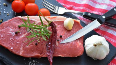 En rå köttbit ligger på en skärbräda. På köttbiten ligger en kryddkvist och en torkad chili. Bredvid köttebiten ligger en kniv, en vitlök och några tomater. Det är strött peppar och salt över köttbiten och skärbrädan.