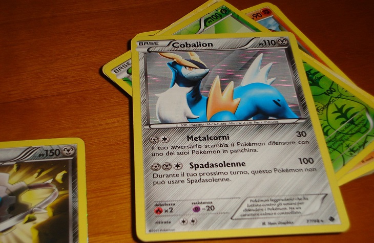 Några pokemonkort ligger i en hög på ett bord. Det översta kortet har gul kant och grå bakgrund. Längst upp är en bild på en blå pokémon med horn. På nedre delen av kortet står text och olika symboler.