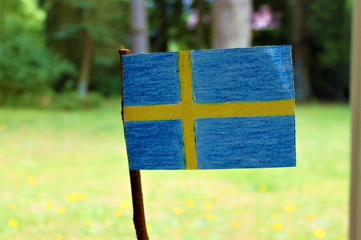 En svensk flagga är ritad med kritor och utklippt. Flaggan sitter på en träpinne. I bakgrundens är det en solig gräsmatta.