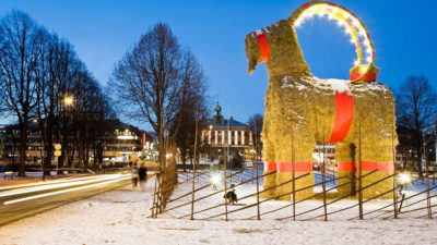 En stor bock av halm står bredvid flera träd. Bocken har röda band runt magen och benen. Runt bocken är det ett staket. På marken är det snö.