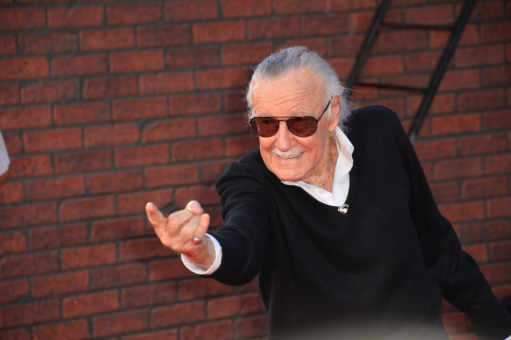 Stan Lee står och pekar framåt med ett finger. Han har grått hår som är bakåtkammat, mörka glasögon och en svart tröja.