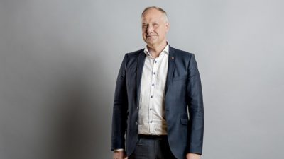Jonas står framför en vit vägg. Han har kostym och skjorta på sig. Han har kort grått hår och ser allvarlig ut.