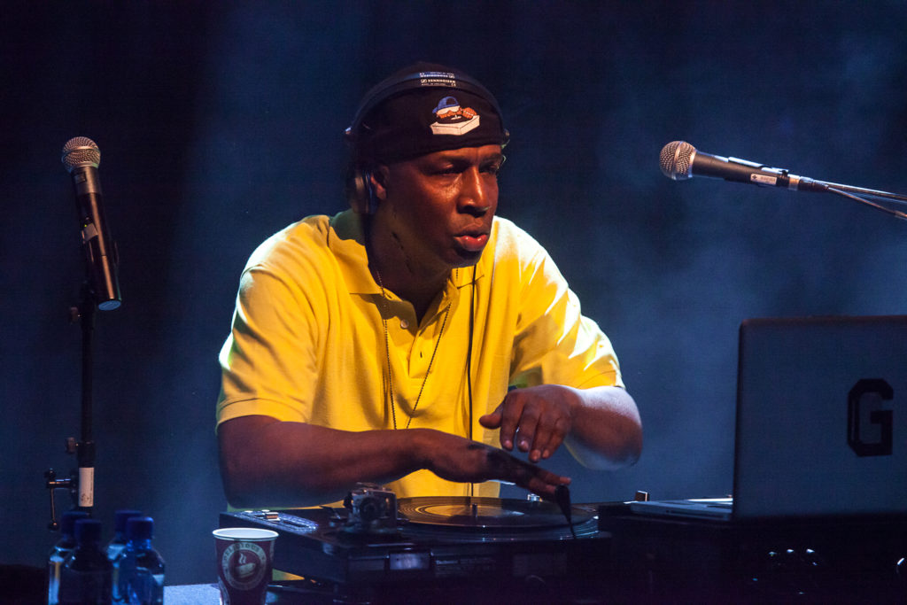 Grandmaster Flash står i ett dj-bås. Han har en backochframvänd keps och stor t-shirt. På bordet framför sig har han dj-utrustning som han spelar med.