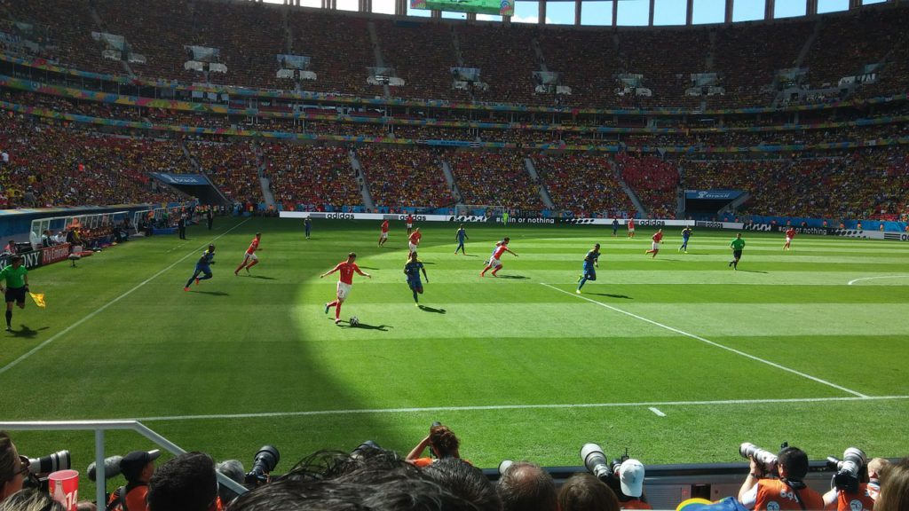 Bild på en fotbollsmatch som är i full gång. Solen lyser mot det perfekt gröna gräset och arenan är fullsatt.