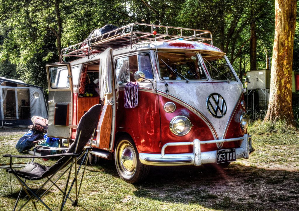 En volkswagenbuss står i ett skogsbryn. Dörren är öppen på sidan och där inne syns möbler. Utanför står några stolar och det hänger kläder på dörrarna.