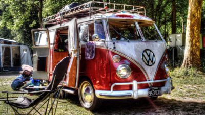 En volkswagenbuss står i ett skogsbryn. Dörren är öppen på sidan och där inne syns möbler. Utanför står några stolar och det hänger kläder på dörrarna.